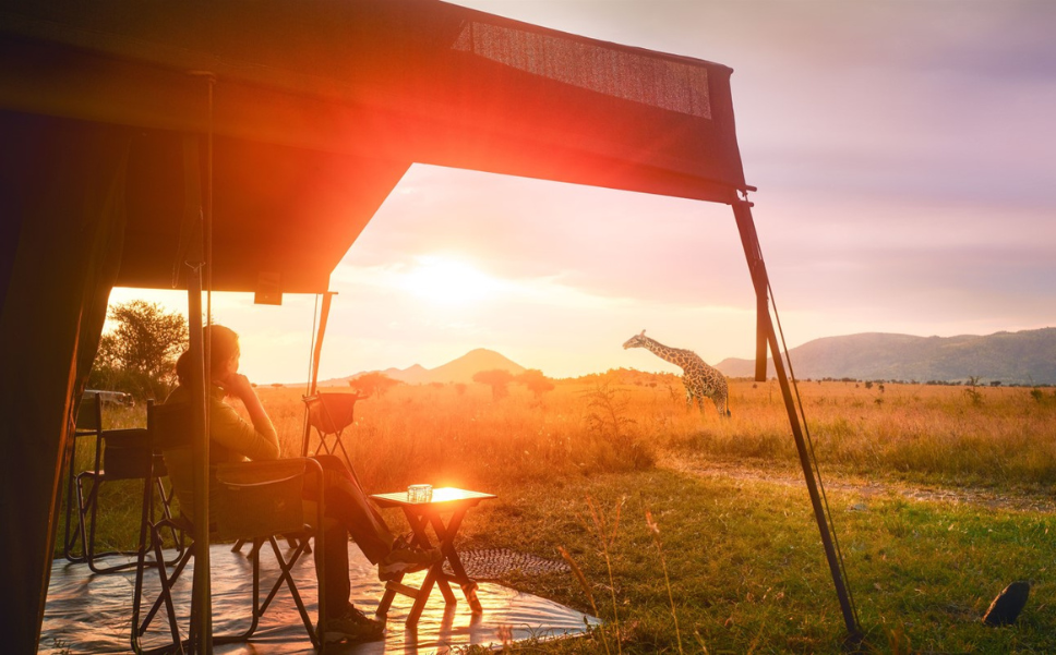 Guest sits outside their tented camp and watched the African wilderness with a giraffe in the foreground.